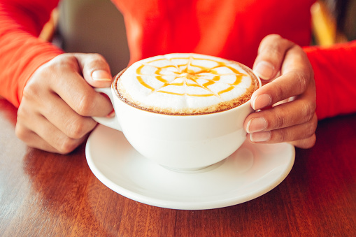 Unrecognizable woman enjoying coffee. Close-up of female hands holding cup with cappuccino. She sitting at table. Cappuccino lover concept