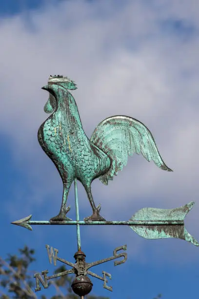 Big cockerel on a weathervane. Metal weather cock pointing north west. Traditional blue green windvane. Weathercock against sky.