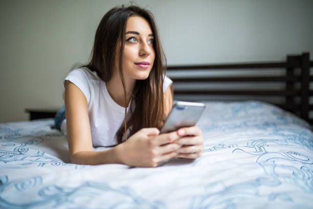 feliz mulher sentada na cama lendo a mensagem de texto no smartphone. sorrindo a garota sentada no quarto em casual em conversa com um amigo numa sala de chat. mulher sorridente surf líquido com o celular em casa. - hotel domestic room telephone technology - fotografias e filmes do acervo