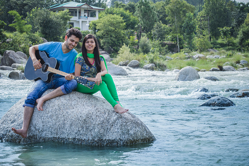 Outdoor image of happy Asian, Indian young couple enjoying their city break and having fun in fresh air. They are sitting on rock in river and playing guitar. Two people, full length and horizontal composition with copy space.
