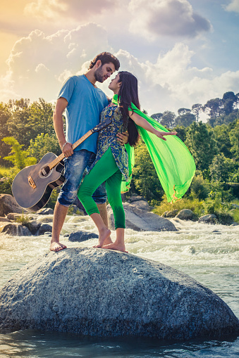 Outdoor image of happy Asian, Indian young couple enjoying their city break and doing romance in fresh air. They are embracing to each other while standing on rock in river holding guitar. Two people, full length and vertical composition with copy space.