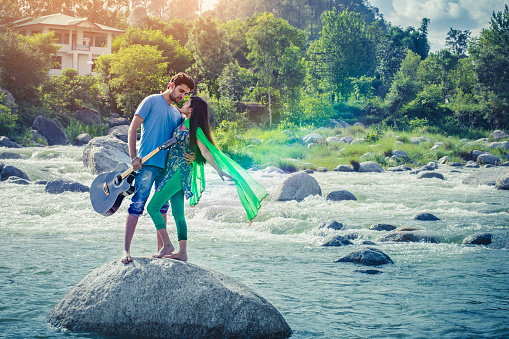 Outdoor image of happy Asian, Indian young couple enjoying their city break and doing romance in fresh air. They are embracing to each other while standing on rock in river holding guitar. Two people, full length and horizontal composition with copy space.