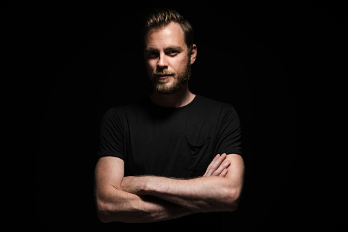 Portrait of a bearded man with an intense look standing in a dark room against a black background.