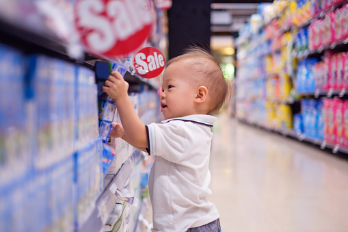 Cute little Asian 18 months / 1 year old toddler baby boy child standing and choosing milk product in grocery store / department store, kid purchasing milk in shop