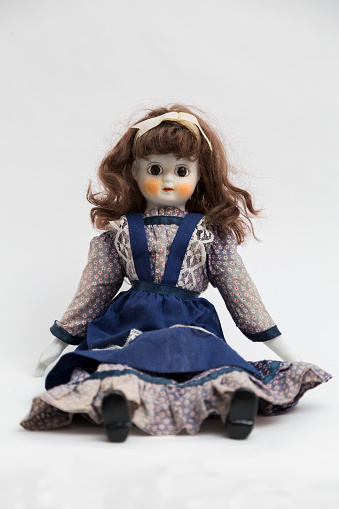 Portrait of ceramic porcelain handmade vintage doll with big eyes, wavy brown hair in old blue textile dress with embroidery, in shirt with gentle floral print , bow, black shoes on white background.