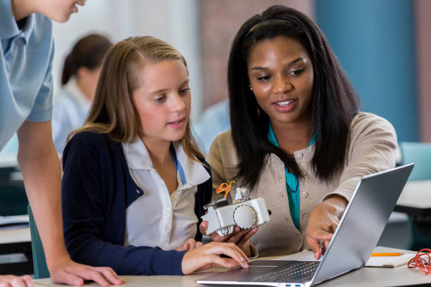 Student works on laptop with teacher in robotics lab A young female middle school student sits at a table with her African American teacher and works on a laptop in the robotics lab. Her teacher holds a robot and points to the computer screen as an unrecognizable male student stands and looks on. female role model stock pictures, royalty-free photos & images