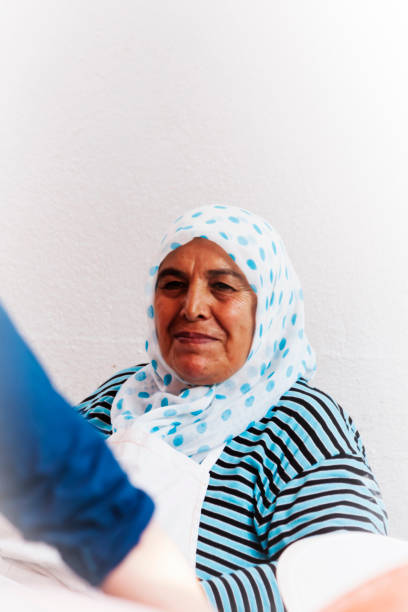 An old turkish woman, wearing traditional white scarf with blue points, is smiling nicely while sitting near a white wall, and chatting with another woman - not visible in the picture. Teos - Seferihisar (Izmir - Turkey). 20 august 2017