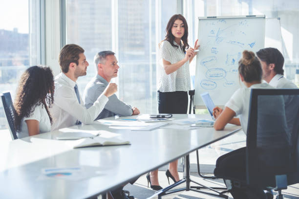 Woman giving a presentation to her team. Woman giving a presentation to her team. She is using a whiteboard with charts and graphs. She is talking. They are in an office boardroom at the table with laptop computers and paperwork. She is smiling. Men and women in the group. course sales stock pictures, royalty-free photos & images