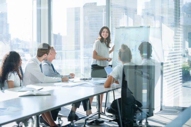 Woman giving a presentation to her team. Woman giving a presentation to her team. She is using a whiteboard with charts and graphs. She is talking. They are in an office boardroom at the table with laptop computers and paperwork. She is smiling. Men and women in the group. business consultation stock pictures, royalty-free photos & images