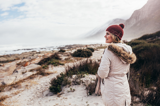 Portrait of beautiful young woman smiling in winter clothes and looking away at the beach. Caucasian female on winter holiday at the sea shore.