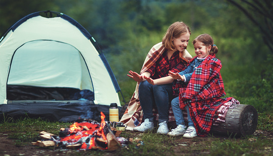 Family mother and child daughter warm their hands by bonfire on  camping trip with a tent