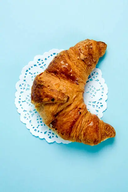 a french croissant on a doily lace paper. Pastel blue background. Minimal color still life photography.Minimal color still life photography Minimal color still life photographM