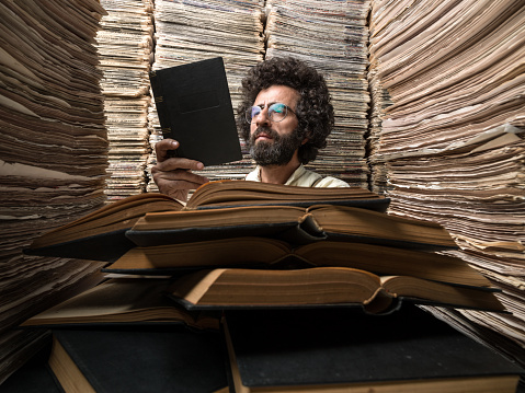 Adult man with dark hair reading book for research in archive of printed media full of large stack of newspapers.The model is wearing a gray shirt, holding hardcover book in hand.Huge amount of real newspapers are used on the desk and on the background.The image was shot in horizontal composition and toned for old fashioned look.