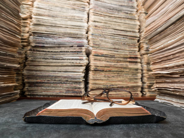 View of large amount of documents, newspapers, and books in classic library. Reading glasses are seen on top of an open book on desk. The book is bible. No people are seen in frame. Shot with a medium format camera.