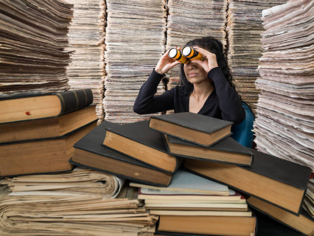 Young mixed race woman doing research in library of media full of newspapers, books and magazines.She is looking through binoculars.The model is wearing a black sweater, holding a hard cover book and binoculars in right hand.The page seen are blank and grunge brown.Huge amount of real newspapers are used.Horizontal framing.