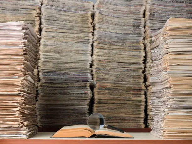 View of large amount of documents, newspapers, and books in classic library. A magnifying glass seen on top of an open book on desk. No people are seen in frame. Shot with a medium format camera.