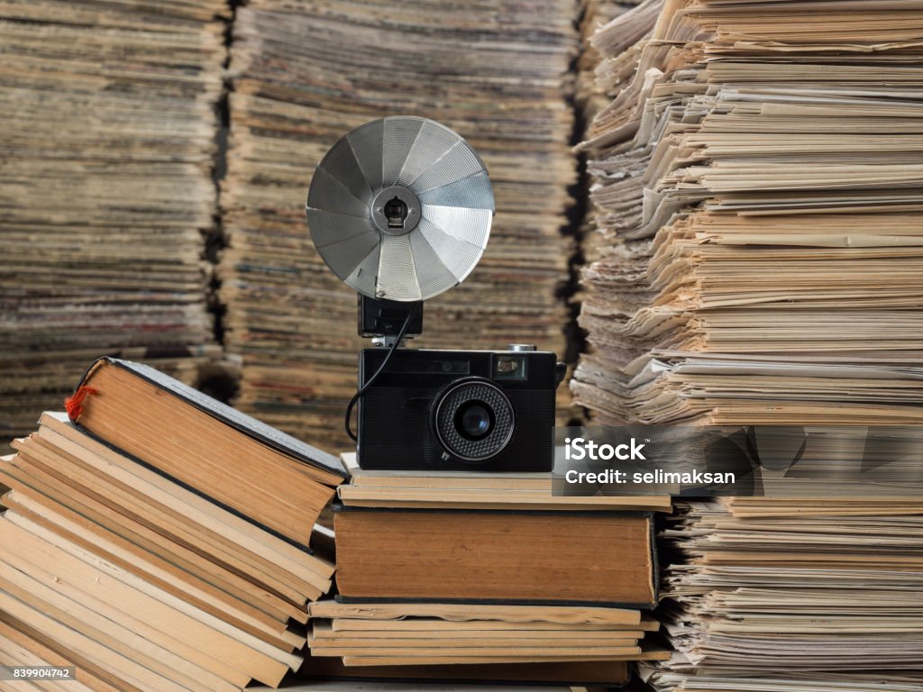 Camera Seen On Large Amount Of Documents, Newspapers, Magazines And Books Media library with large amount of newspapers, magazines, books and documents. An old fashioned camera with folding flash is seen on top of books. No people are seen in frame. Shot with a medium format camera. Horizontal framing. Camera - Photographic Equipment Stock Photo