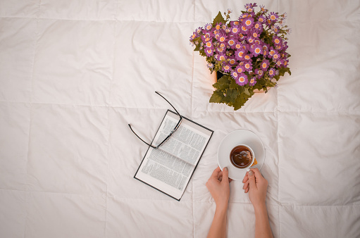 Woman's arm with cup of black tea, glasses, book and purple flowers on white bed.