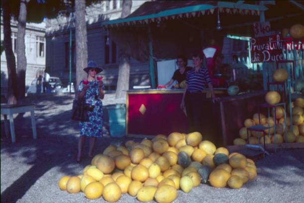 Palermo, Sicily, Italy, 1964. Market square in the center of Palermo. A fruit and vegetable trader offers melons. A tourist poses for photographs next to the melon pile