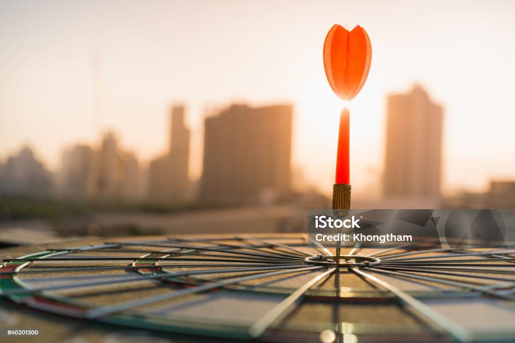 Red dart arrow hitting in the target center of dartboard with modern city and sunset background. Target business, achieve and victory concept . Achievement Stock Photo