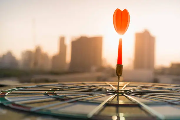 Red dart arrow hitting in the target center of dartboard with modern city and sunset background. Target business, achieve and victory concept .