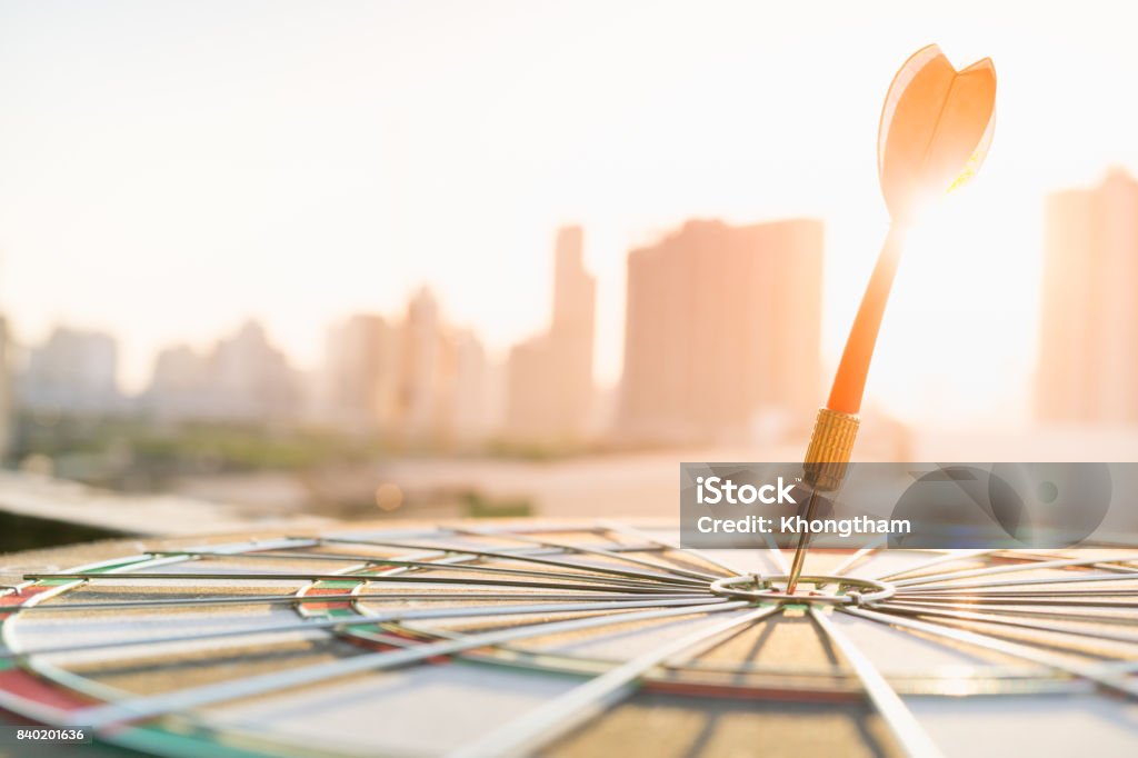 Dardo rojo flecha golpea en el centro de Diana de dardos con la ciudad moderna y fondo puesta de sol. Objetivo negocio, alcanzar y concepto de Victoria. - Foto de stock de Foco - Determinación libre de derechos