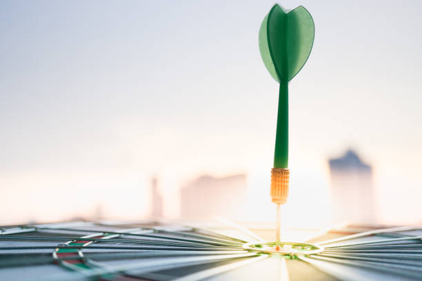 vert dart flèche frappant dans le centre de la cible de jeu de fléchettes avec la ville moderne et fond coucher de soleil. cibler les entreprises, à réaliser et le concept de la victoire. - market target photos et images de collection