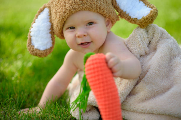 baby in the hat like a bunny with carrot toy on green grass outdoors - ravena imagens e fotografias de stock