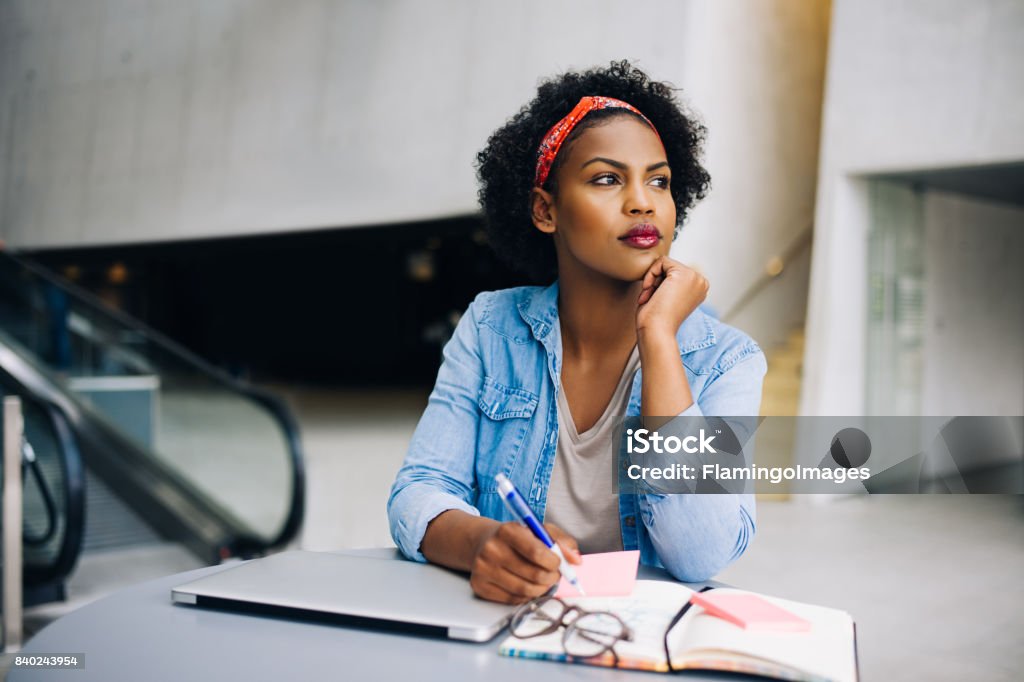 Young African female entrepreneur dreaming up new business ideas Focused young African female entrepreneur deep in thought while working at a table in a modern office building lobby Contemplation Stock Photo