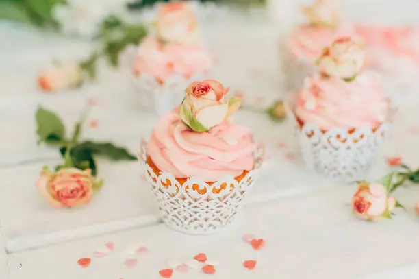 Delicate tasty muffins with a pink cream decorated with real roses and sprinkled with hearts on a wooden background. Homemade food. Toning