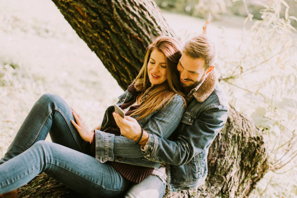 istock Couple in love sitting on a tree in autumn park and using mobile phone 840317034