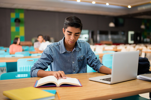 A male college student sitting in the cafeteria with a laptop and reading a book.