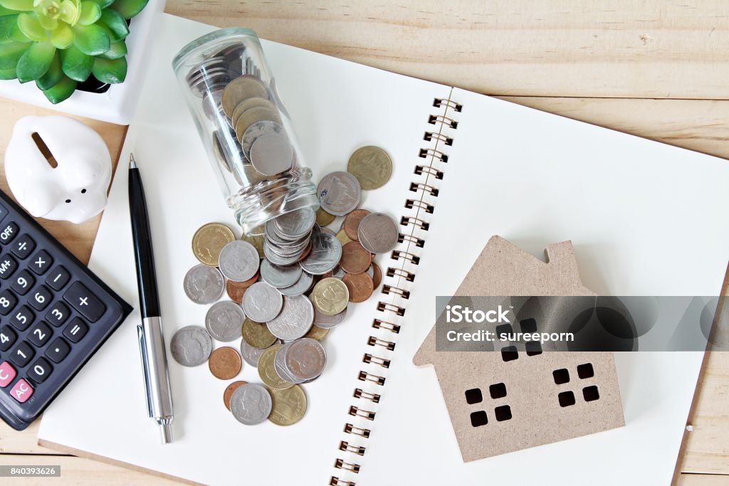 Flat lay or top view of wood house model and coins scattered from glass jar on open blank notebook paper Business, finance, savings, property ladder or mortgage loan concept : Flat lay or top view of wood house model and coins scattered from glass jar on open blank notebook paper House Stock Photo