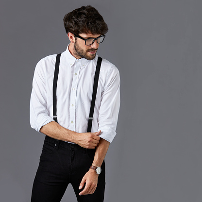 Confident businessman standing against gray background. Handsome male professional is in white shirt and suspenders. Creative executive is looking down while folding sleeve.