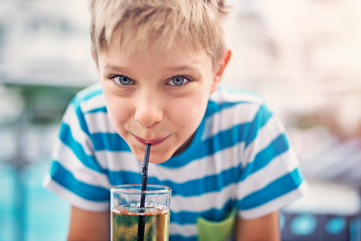 Little boy aged 7 drinking a glass of apple juice. The boy is smiling at the camera while sipping the juice.
