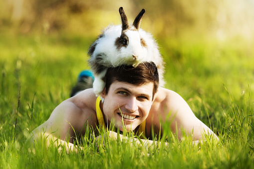 A smiling and muscular young man lies in long grass with his pet angora rabbit rather bizarrely but happily sitting on his head.