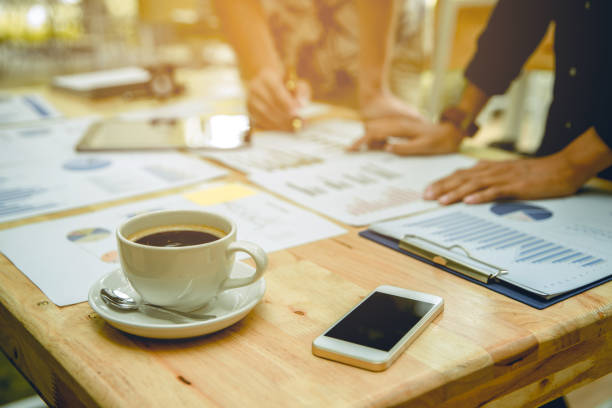 istock Two businessmen working and analyzing graphs papers on wooden table. there is cup of coffee and smartphone on table. Focus at cup of coffee and smart phone. 840474660