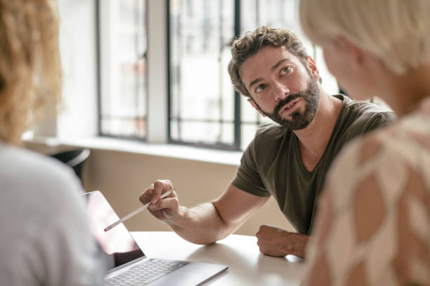 Mature businessman with beard pointing at laptop with pencil and listening to colleague Businessman in his 40s with beard in meeting with two people, casual creative professional talking with clients persuasion stock pictures, royalty-free photos & images