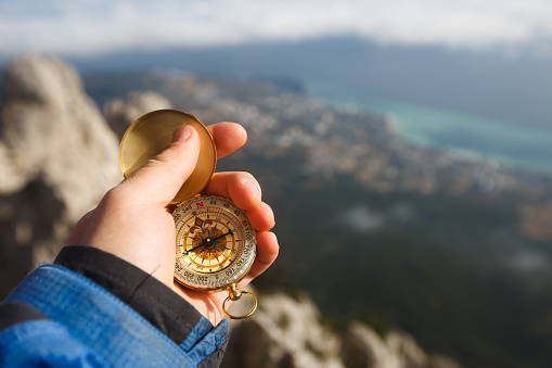 Point of view photo of explorer man searching direction with golden compass in his hand above clouds with autumn mountains and sea background. Navigation and travel concept