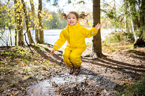 Cute little girl jumping in muddy puddle wearing yellow rubber overalls. Happy childhood. Sunny autumn forest