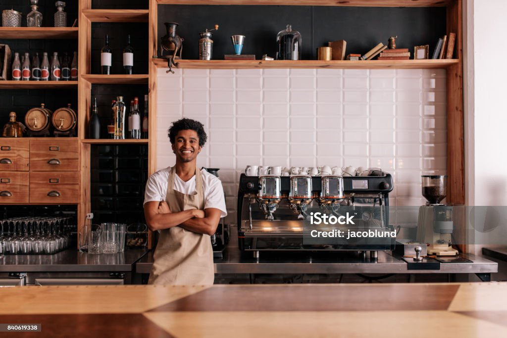 Barista in apron looking at camera and smiling Portrait of confident young man wearing apron standing behind cafe counter. Barista in apron looking at camera and smiling. Coffee Shop Stock Photo