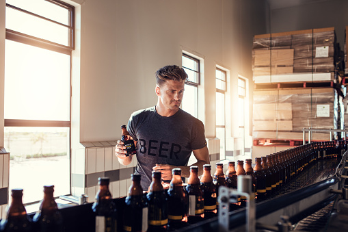 Young man working at small craft beer making factory and checking the quality of beer. Young businessman testing the beer bottle at brewery.