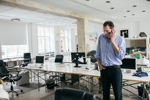 A manager is talking on his smartphone in an empty office space after his employees have left at the end of the day.