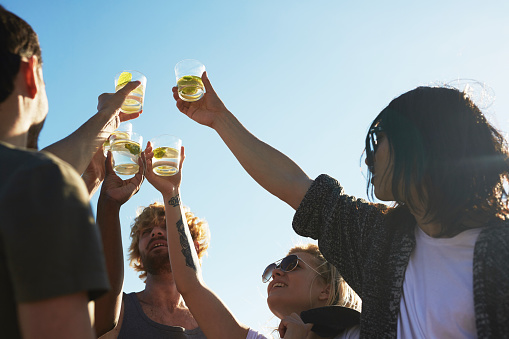 Low angle view of happy young friends toasting with mojito cocktails while having fun at outdoor party, cloudless blue sky on background