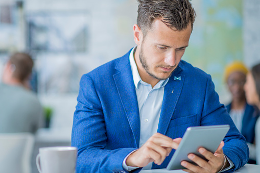 A multi-ethnic group of adults are working indoors in an office. They are wearing formal clothing. A Caucasian man is working on a tablet computer while his coworkers talk in the background.