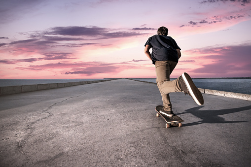 Skateboarder pushing on a concrete pavement along the harbour during the sunset.
