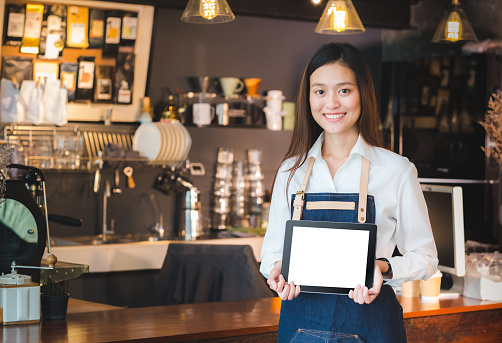 Sonriente mujer asiática barista con cálculo de tablet en blanco frente a cafetería bar de contador, se burlan de un espacio para la pantalla de menú o de diseño, trazado de recorte en pantalla. photo
