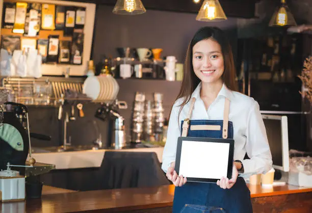 Photo of Smiling asian woman barista holding blank tablet compute in front of coffee shop counter bar,Mock up space for display of menu or design,clipping path on screen.