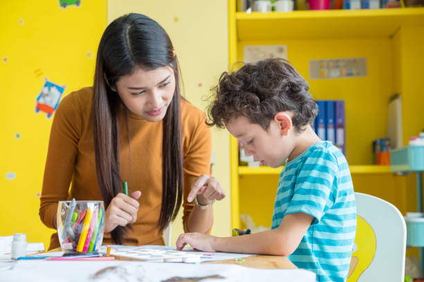 Asian woman teacher teaching boy kid to paint color book on table in classroom,kindergarten education school Asian woman teacher teaching boy kid to paint color book on table in classroom,kindergarten education school. asian nanny stock pictures, royalty-free photos & images