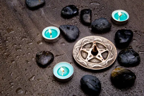 Photo of Incense burning in silver pentagram with blue candles and black rocks on gray slate stone background